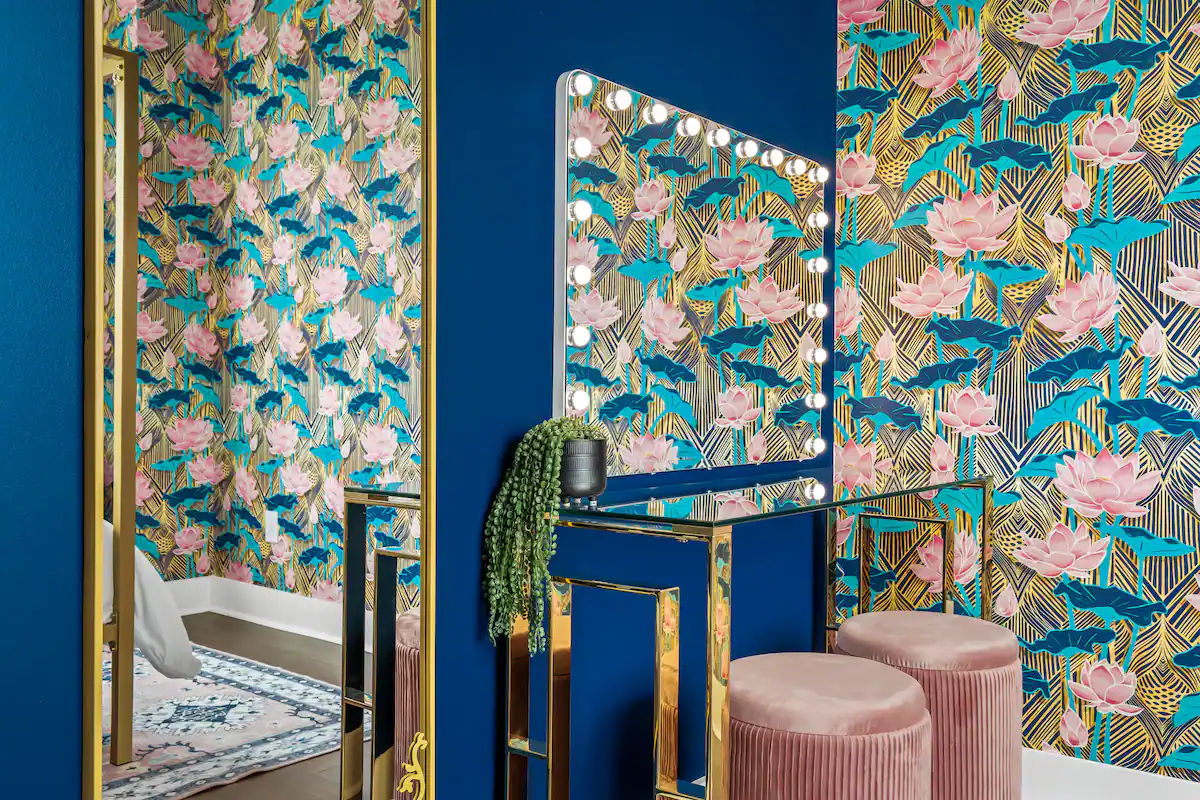 Vibrant vanity nook with lit Hollywood mirror, deep blue wall, gold-framed glass table, bold pink lotus wallpaper, two pink velvet stools and trailing plant
