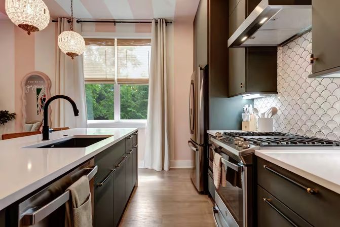 Bright modern kitchen with central island, matte black cabinets, white quartz countertops, stainless steel range and fridge, scallop-pattern tile backsplash, hanging globe pendants, and a window overlooking leafy green trees.