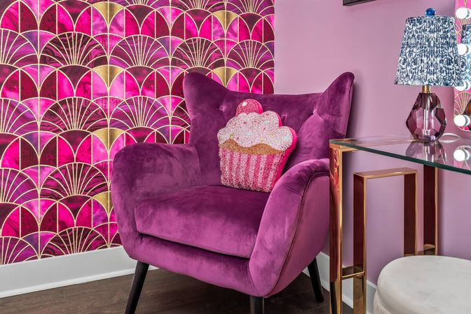 Plush magenta velvet armchair with a sparkly cupcake pillow next to a glass-and-gold vanity lamp, set against fuchsia and gold Art Deco wallpaper.