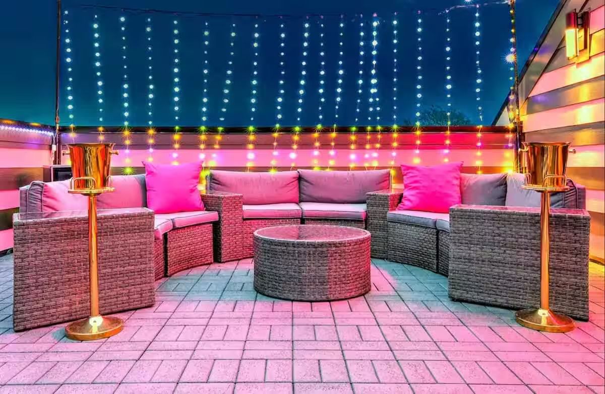 Nighttime urban rooftop patio with curved wicker sectional, pink throw pillows, round wicker coffee table, hanging string lights and gold champagne buckets on stands.