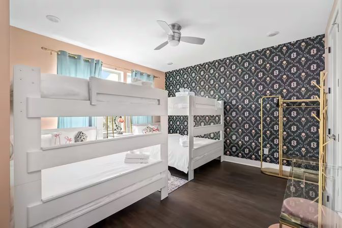 Modern kids' bedroom with two white twin bunk beds, dark floral accent wallpaper, turquoise curtains, ceiling fan, gold clothing rack, and dark hardwood floors