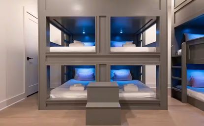 Sleek modern gray built-in bunk room with four roomy bunk pods, blue LED ambient lighting, white linens and folded towels, and a central step leading to the lower beds — cozy shared sleeping space.