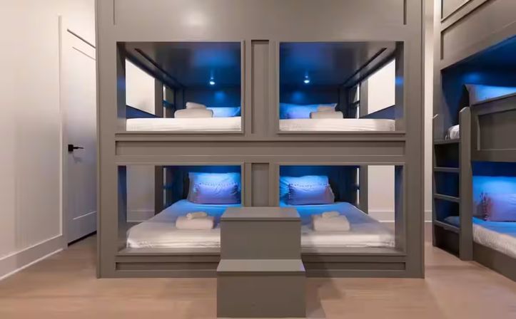 Sleek modern gray built-in bunk room with four roomy bunk pods, blue LED ambient lighting, white linens and folded towels, and a central step leading to the lower beds — cozy shared sleeping space.