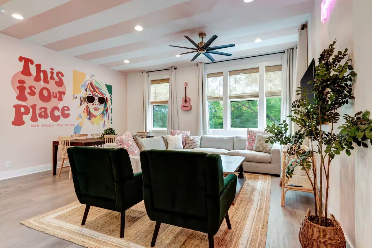Sunlit modern living room with a playful mural reading "This is our Place", light gray sectional sofa, two dark green armchairs, large windows with a leafy view, ceiling fan, pink guitar, dining table and potted plants.