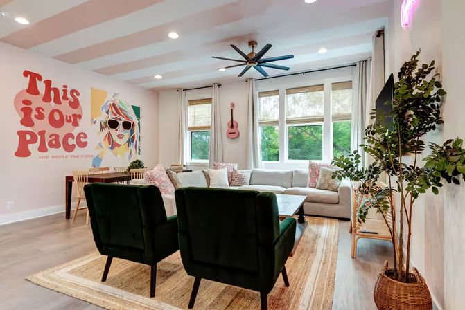 Sunlit modern living room with a playful mural reading "This is our Place", light gray sectional sofa, two dark green armchairs, large windows with a leafy view, ceiling fan, pink guitar, dining table and potted plants.