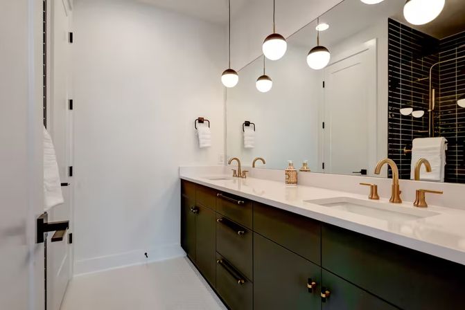 Sleek modern bathroom with a double white quartz vanity, dark green lower cabinets, brass-gold faucets and globe pendant lights, large mirror reflecting a black subway-tile shower.