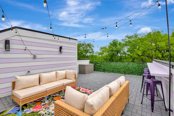 Modern rooftop patio lounge with wicker sofas and cream cushions on a colorful floral rug, purple bar stools at a counter, overhead string lights, green privacy hedge and treetop views under a blue sky.