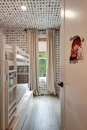 Playful cat-themed kids' bedroom with repeating cat-face wallpaper on walls and ceiling, white bunk bed with trundle, beige curtains framing a window overlooking green trees, light hardwood floor, and a door decal of a woman holding a cat reading "Karma is a cat."