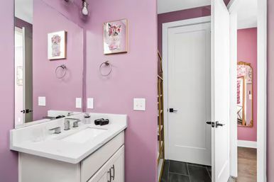 Cheerful pink powder room with white single-sink vanity, large mirror, chrome faucet, towel ring, floral wall art, and an open white door to an adjoining room.
