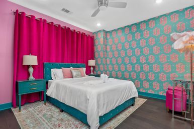 Playful colorful bedroom interior with a teal upholstered bed and nightstands, crisp white bedding, bold fuchsia floor-to-ceiling curtains, teal-and-pink patterned wallpaper, matching lamps, feather floor lamp, and modern ceiling fan.