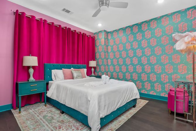 Playful colorful bedroom interior with a teal upholstered bed and nightstands, crisp white bedding, bold fuchsia floor-to-ceiling curtains, teal-and-pink patterned wallpaper, matching lamps, feather floor lamp, and modern ceiling fan.