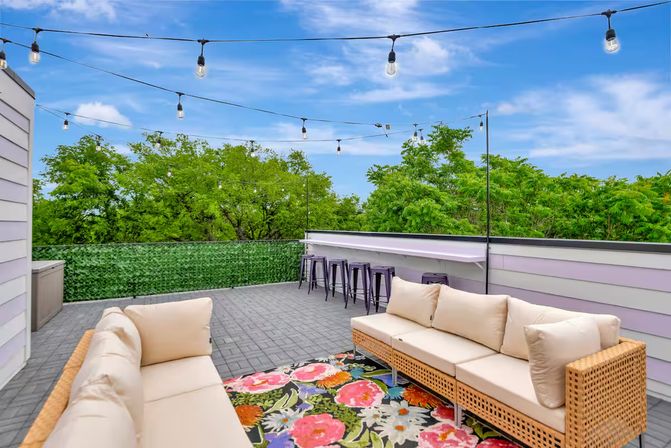 Rooftop patio lounge with wicker sofas and beige cushions on a colorful floral rug, bar stools along the railing, string lights overhead and leafy treetop views under a bright blue sky.