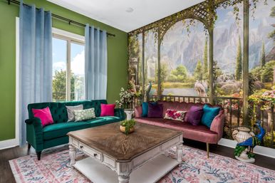 Eclectic living room interior with green walls and pale-blue curtains, teal velvet sofa and mauve velvet loveseat with bright pillows, large scenic wall mural of a mountain lake garden with peacocks, rustic wood coffee table on a floral rug.