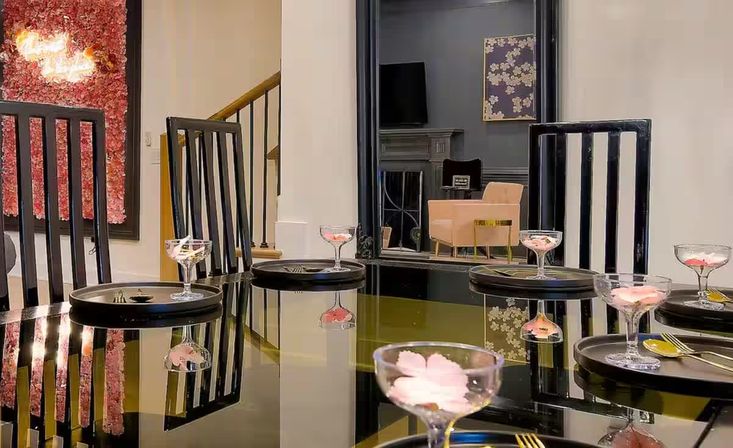 Contemporary indoor dining room with glossy black table reflecting coupe glasses filled with pink flower petals, black slat-back chairs, round black chargers and gold flatware, floral wall art and a cozy lounge chair in the background.