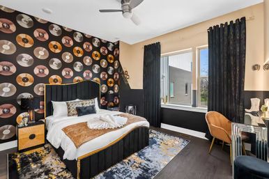 Modern bedroom interior with vinyl-record accent wall, black upholstered bed with gold trim, white bedding and tan throw, patterned rug, black curtains, window and orange accent chair.