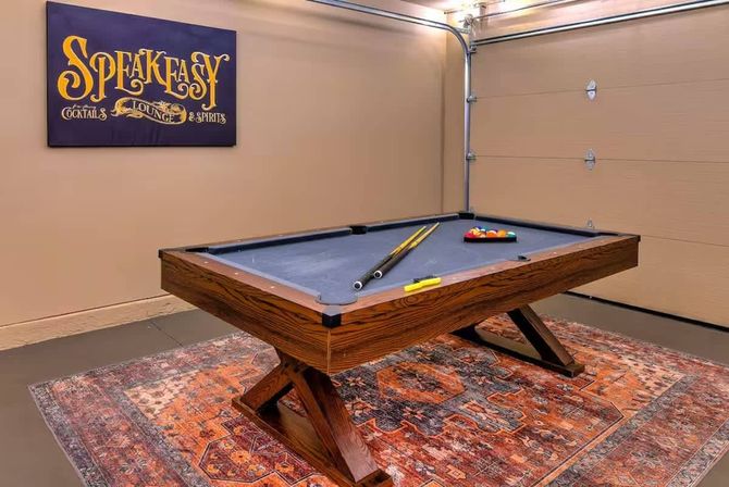 Wooden pool table with gray felt in a cozy garage game room on a patterned vintage rug, cues and racked balls on the table and a decorative "Speakeasy" sign on the wall.