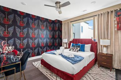 Rock-themed modern bedroom with red velvet bed, blue accent pillows and throw, winged-guitar wallpaper, wooden nightstand, desk with neon music sign and large window