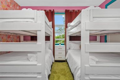 Bright family-friendly kids' bunk room with white bunk beds, pink accent walls and patterned wallpaper, green shag runner, small white dresser under a curtained window with a leafy view.