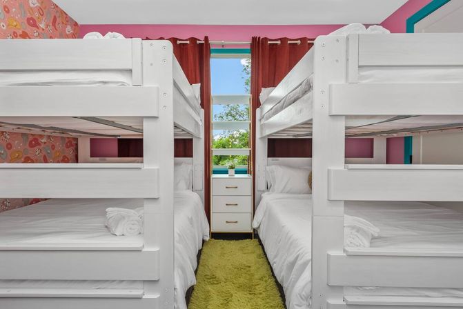 Bright family-friendly kids' bunk room with white bunk beds, pink accent walls and patterned wallpaper, green shag runner, small white dresser under a curtained window with a leafy view.