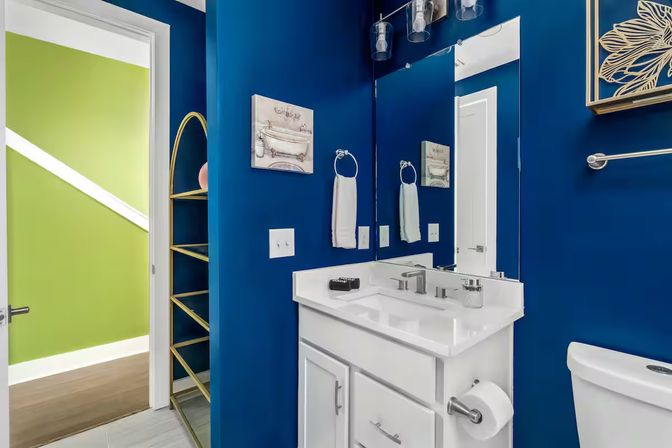 Modern residential bathroom with deep navy blue walls, white vanity and sink, large mirror and light fixture, gold arched shelving, towel ring and toilet, bright green hallway visible through open door.