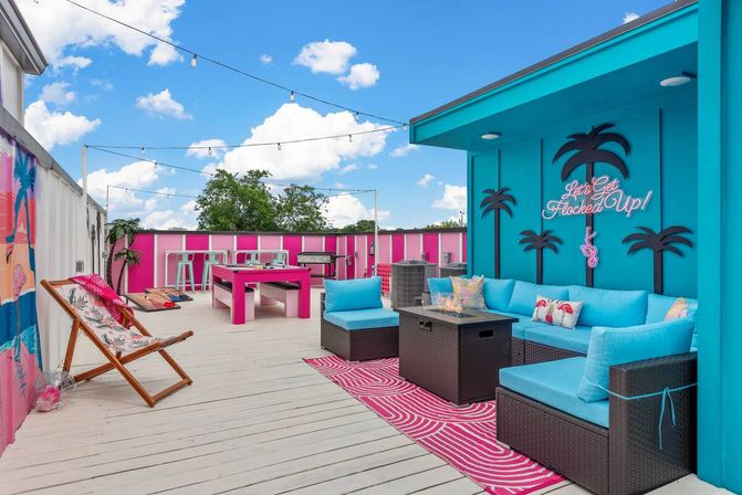 Colorful rooftop patio and urban deck with turquoise sectional, pink picnic table, neon sign and palm silhouettes, string lights, cornhole and summer lounge seating.