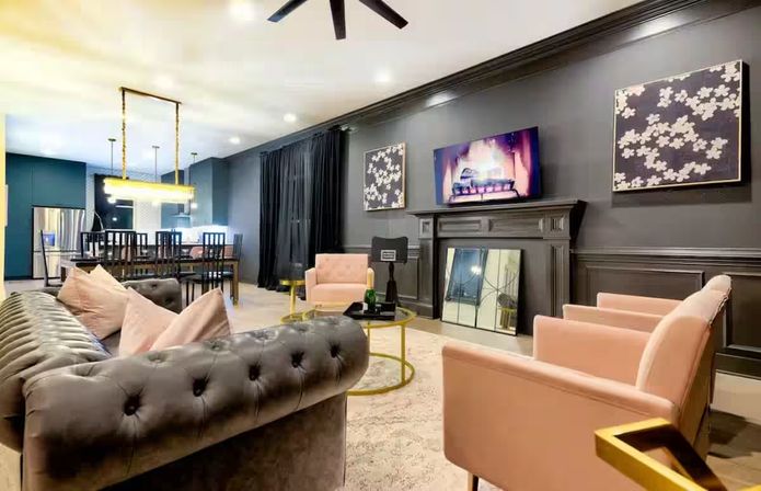 Stylish open-plan city apartment living room with tufted gray sofa, blush-pink armchairs, gold-accent round coffee table, dark paneled fireplace wall with TV, and adjacent dining area with pendant lights and stainless-steel fridge.