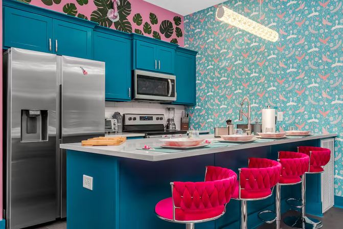 Vibrant modern kitchen with teal cabinets and island, stainless steel appliances, bright pink velvet bar stools, tropical flamingo and monstera wallpaper, and place settings on the countertop.