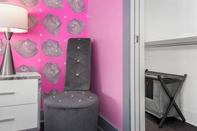 Pink bedroom corner with cowboy-hat patterned wallpaper, mirrored white nightstand and silver lamp, plush gray tufted vanity chair with crystal buttons, and an open closet with a gray laundry hamper.