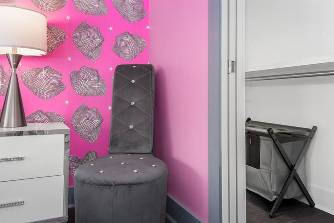 Pink bedroom corner with cowboy-hat patterned wallpaper, mirrored white nightstand and silver lamp, plush gray tufted vanity chair with crystal buttons, and an open closet with a gray laundry hamper.