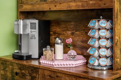 Cozy coffee station on rustic wooden shelf with a silver single-serve coffee maker, pink tray holding sugar packets and a milk jar, small vase with a pink rose, and a rotating rack of donut-labeled coffee pods.