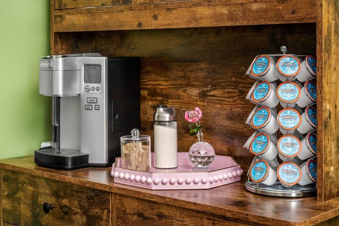 Cozy coffee station on rustic wooden shelf with a silver single-serve coffee maker, pink tray holding sugar packets and a milk jar, small vase with a pink rose, and a rotating rack of donut-labeled coffee pods.