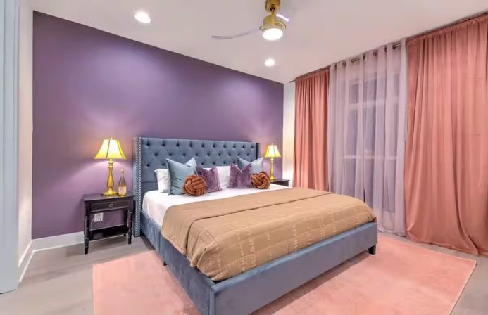 Modern cozy bedroom with a purple accent wall, tufted blue-gray king bed, beige quilted comforter, decorative pillows and knot cushions, gold bedside lamps, pink sheer curtains and plush rug.