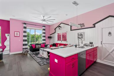 Open-concept living room and kitchen with bright hot-pink island and accent walls, black-and-white striped curtains, tufted black sofa, farmhouse barn door and large window with tree views.