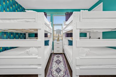 Bright vacation-rental bunk room with two white twin-over-twin bunk beds facing each other, teal accent walls and blue-gold patterned wallpaper, center window above a white three-drawer nightstand with lamp, purple curtains, teal throw pillows and a patterned runner rug.