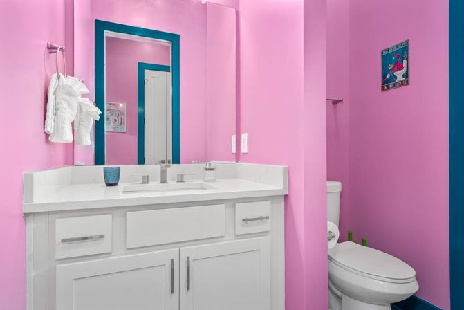 Playful bright pink powder room with white vanity and countertop, teal-framed mirror, folded white towels and a toilet with flamingo wall art.
