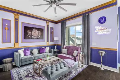 Glam purple-and-gold urban apartment living room with velvet tufted sofas, mirrored coffee table, chandelier wall art, neon sign reading 'Too glam to give a damn', balcony window and decorative gold trim.