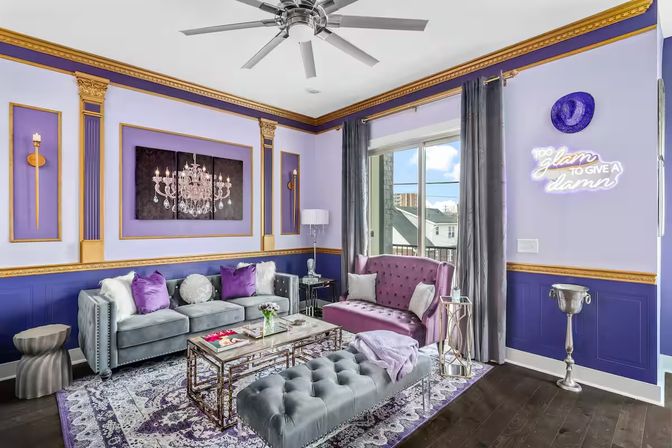 Glam purple-and-gold urban apartment living room with velvet tufted sofas, mirrored coffee table, chandelier wall art, neon sign reading 'Too glam to give a damn', balcony window and decorative gold trim.