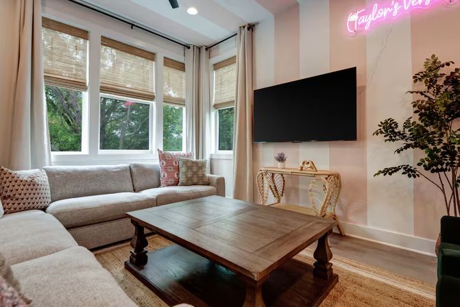 Bright cozy living room with L-shaped gray sectional, wooden coffee table, large windows with bamboo shades and tree view, wall-mounted TV, pink neon wall accent, and potted plant