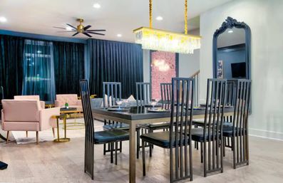 Chic modern open-concept dining and living area with black high-back chairs around a glass-top table, crystal chandelier, blush-pink armchairs, floor-to-ceiling dark curtains, large decorative mirror, and light wood floors.