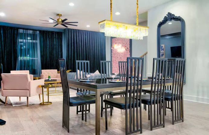 Chic modern open-concept dining and living area with black high-back chairs around a glass-top table, crystal chandelier, blush-pink armchairs, floor-to-ceiling dark curtains, large decorative mirror, and light wood floors.
