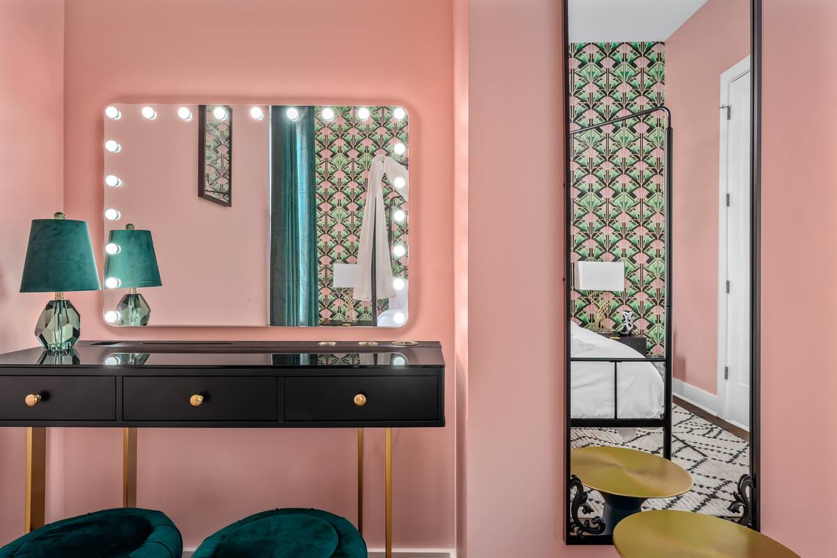 Pink dressing area with black vanity and gold accents, lightbulb-framed Hollywood mirror, teal velvet lamps and stools, and art-deco green patterned wallpaper reflecting a bed.