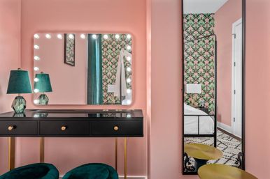 Pink dressing area with black vanity and gold accents, lightbulb-framed Hollywood mirror, teal velvet lamps and stools, and art-deco green patterned wallpaper reflecting a bed.