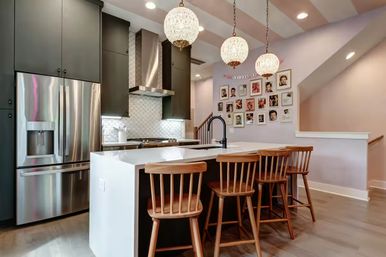 Contemporary open-plan kitchen with white waterfall island, four wooden bar stools, stainless steel appliances, dark cabinets, patterned tile backsplash, three crystal globe pendant lights, framed photo gallery on a pastel wall, and hardwood floors.