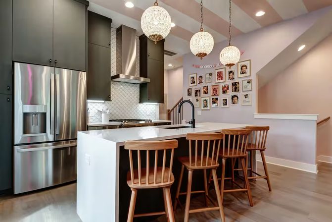Contemporary open-plan kitchen with white waterfall island, four wooden bar stools, stainless steel appliances, dark cabinets, patterned tile backsplash, three crystal globe pendant lights, framed photo gallery on a pastel wall, and hardwood floors.