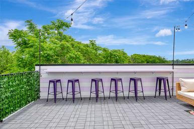 Cheerful rooftop patio with a long bar counter, six purple metal stools, string lights, green hedge railing, wicker sofa, and treetops under a bright blue sky.