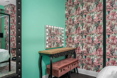 Bedroom vanity nook with turquoise wall and pink cactus-pattern wallpaper, wooden console table with Hollywood-style lightbulb mirror and pink velvet storage bench