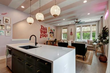 Bright open-concept kitchen and living room with white waterfall island and black faucet, three crystal pendant lights, striped ceiling beams, playful mural reading "This is our place", pink guitar by sunlit windows, neutral sofa, green armchairs and woven rug overlooking a leafy view.