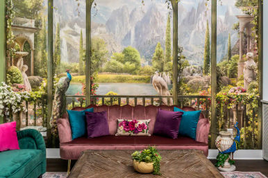 Opulent living room interior with mauve velvet sofa, teal and purple pillows, wooden coffee table and potted plant, backed by a floor-to-ceiling classical garden mural with peacocks, statues, fountains and a mountain lake vista.