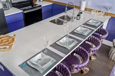 Modern kitchen island with white countertop and stainless double sink, vibrant blue lower cabinets, four purple velvet swivel bar stools with gold bases, four place settings with glass plates and crystal wine glasses, and a decorative "cheers" sign.