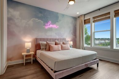 Sunlit modern bedroom with pastel cloud mural, pink velvet bed and blush pillows, neon 'Love' sign above the headboard, bedside lamps and large windows with a neighborhood view.